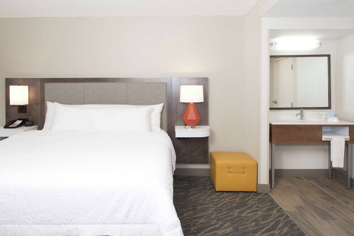 Een hotelkamer met een bed, bedlampjes, een nachtkastje met telefoon, een klein oranje krukje en een badkamer met wastafel en spiegel.