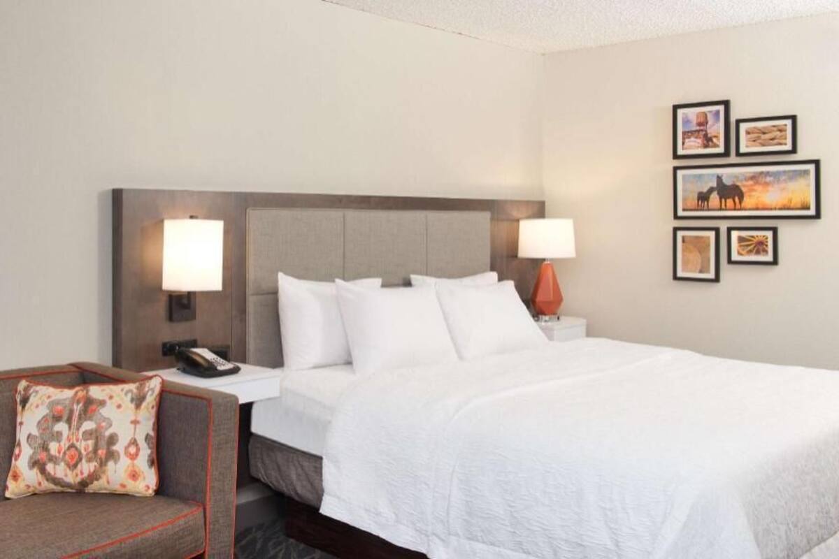 Een hotelkamer met een bed, bedlampjes, een telefoon, een stoel met kussen, en lijstjes aan de muur.