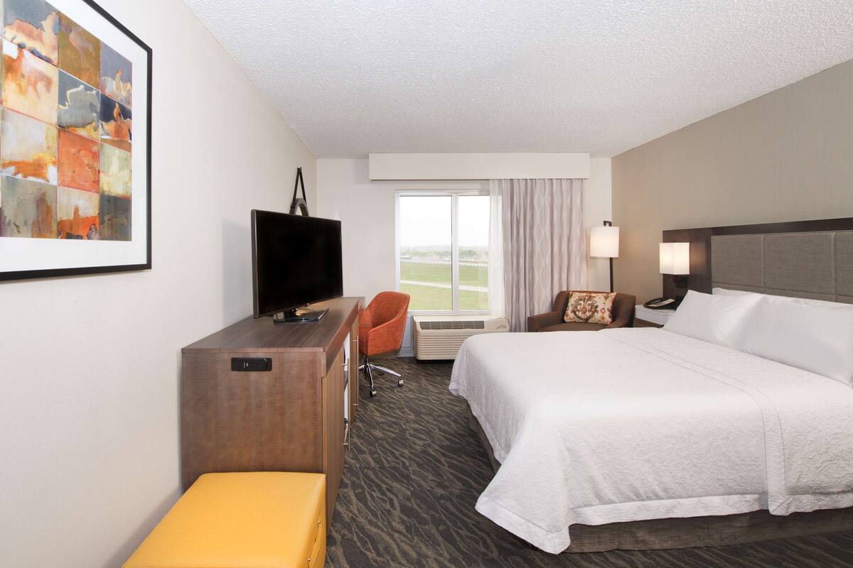Een hotelkamer met een bed, een bureau met een televisie, een stoel, een raam met uitzicht en een schilderij aan de muur.