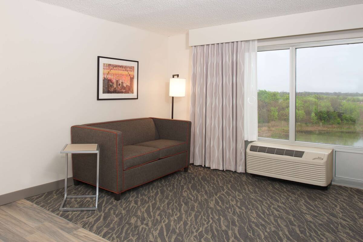 Een hotelkamer met een grijze bank, een klein bijzettafeltje, een staande lamp, een raam met gordijnen en een radiator.