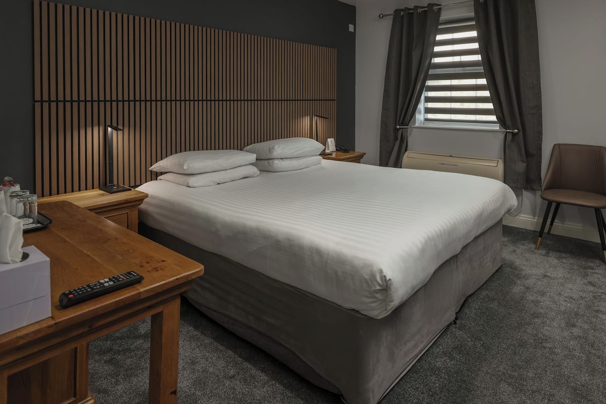 Ein ordentlich bezogenes Bett mit Kissen, ein Nachttisch mit Fernbedienung, eine Lampe und ein Sessel in einem modernen Zimmer, das ein schlaufenförmiger Bettrahmen und ein Fenster mit Vorhängen hat.