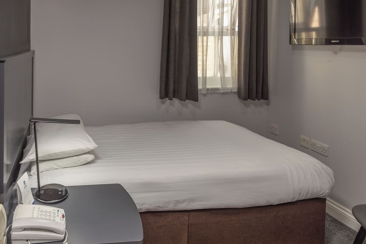 Ein Hotelzimmer mit einem Bett, einem Schreibtisch mit Telefon, einer Lampe und einem an der Wand befestigten Fernseher.