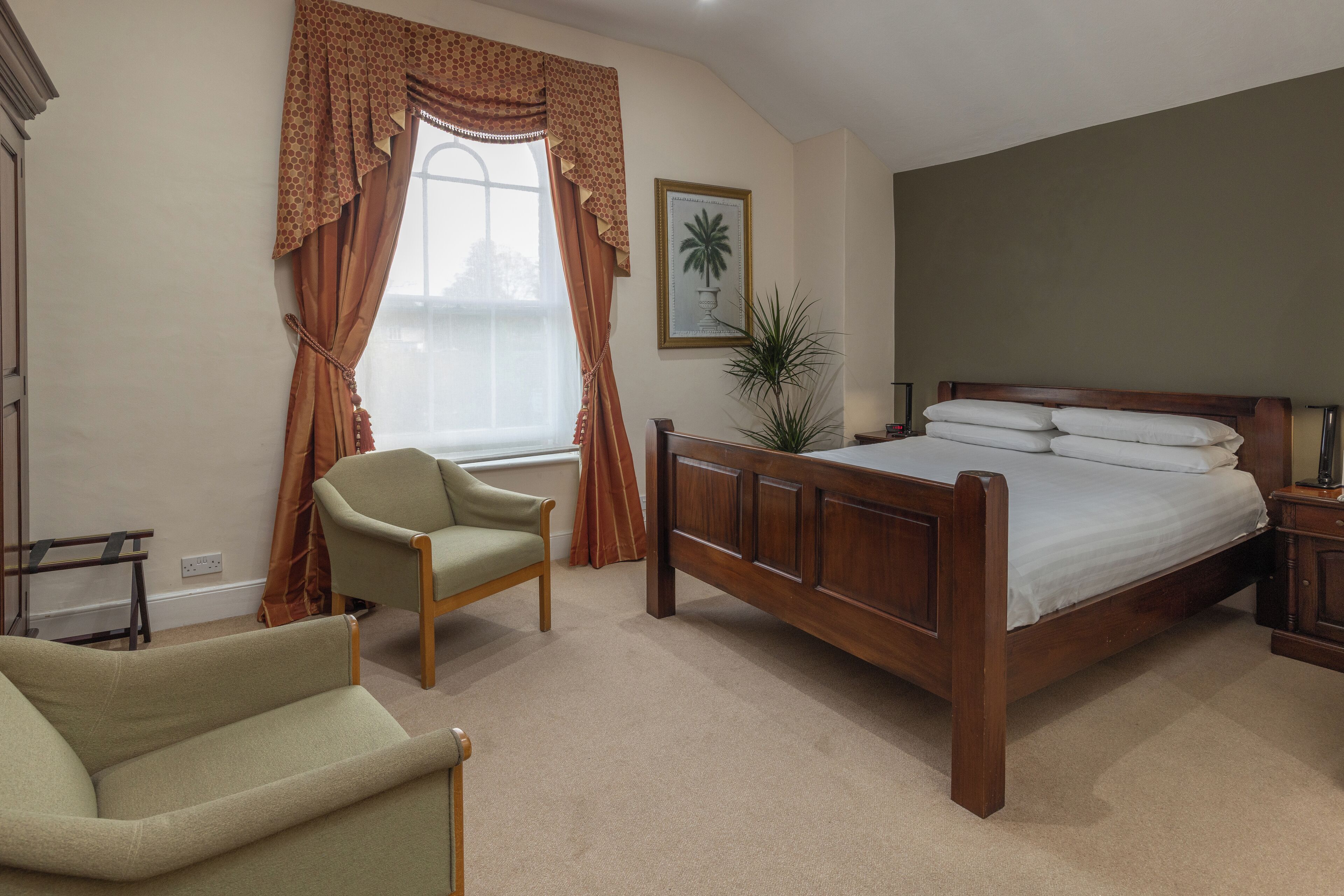 A bedroom with a wooden bed, a green armchair, a window with curtains, and a framed picture on the wall.