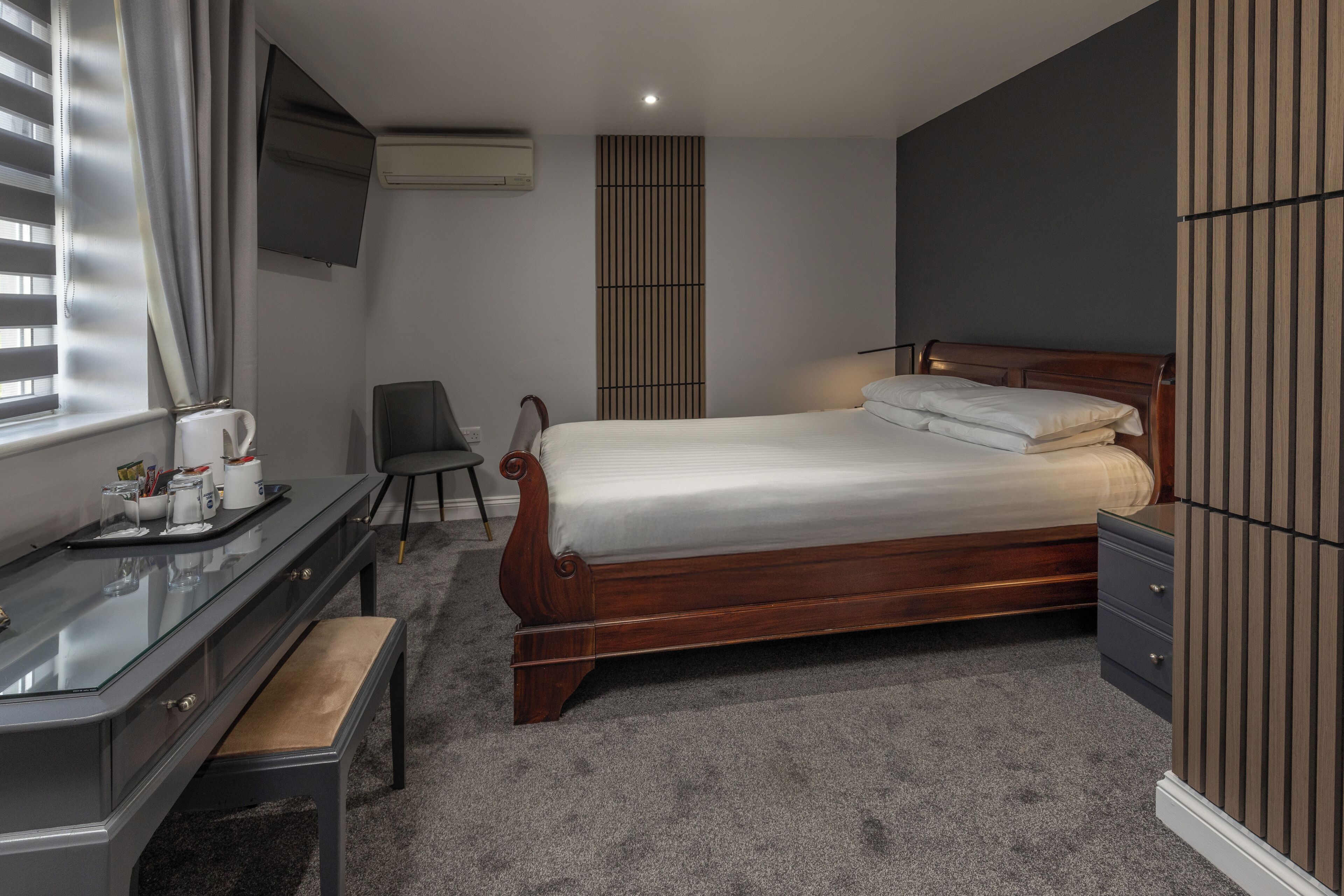 A bedroom with a wooden bed, a desk with a chair, a TV mounted on the wall, and a window with curtains.