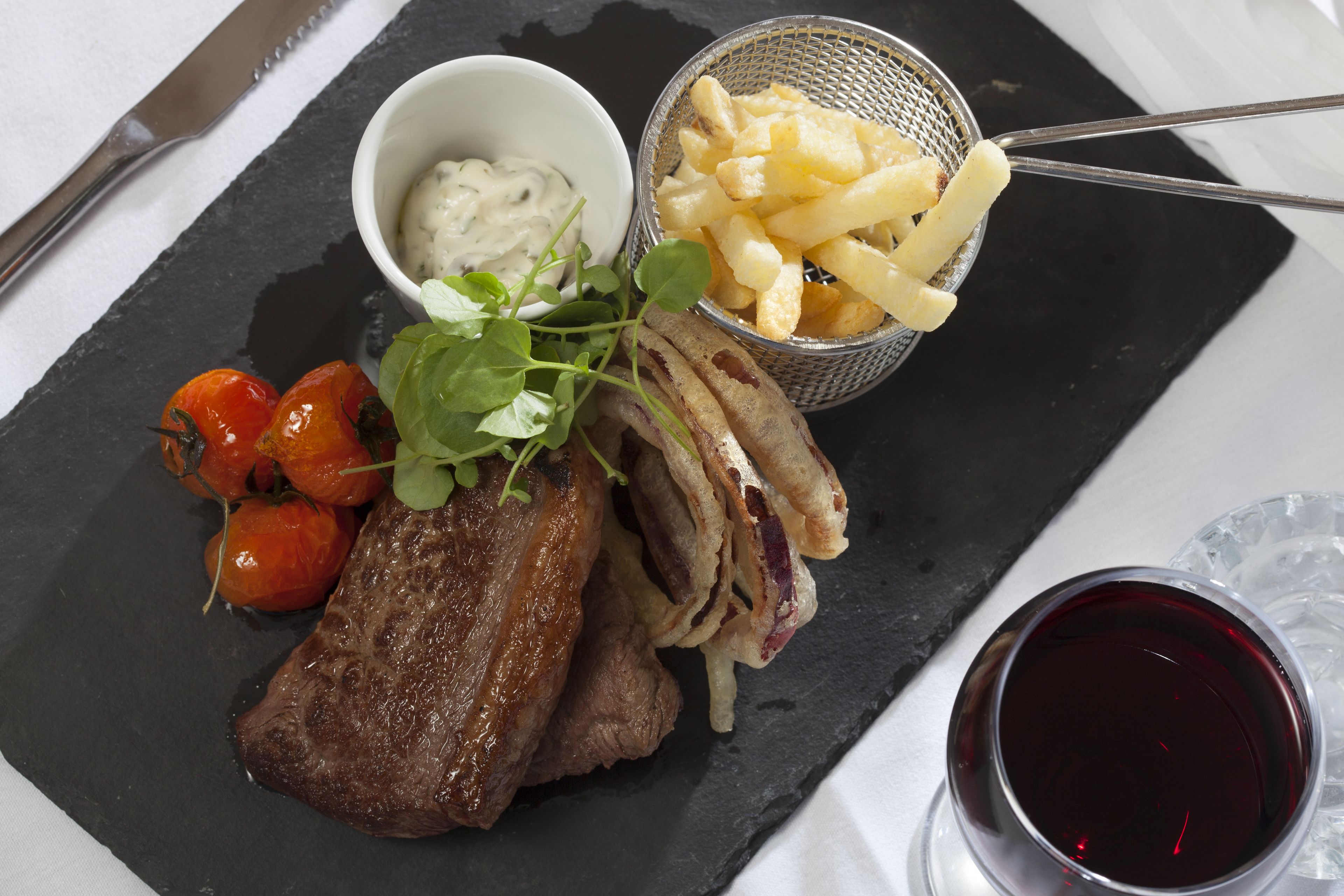 A black slate serving board with a steak, cherry tomatoes, a small bowl of sauce, and a basket of fries.