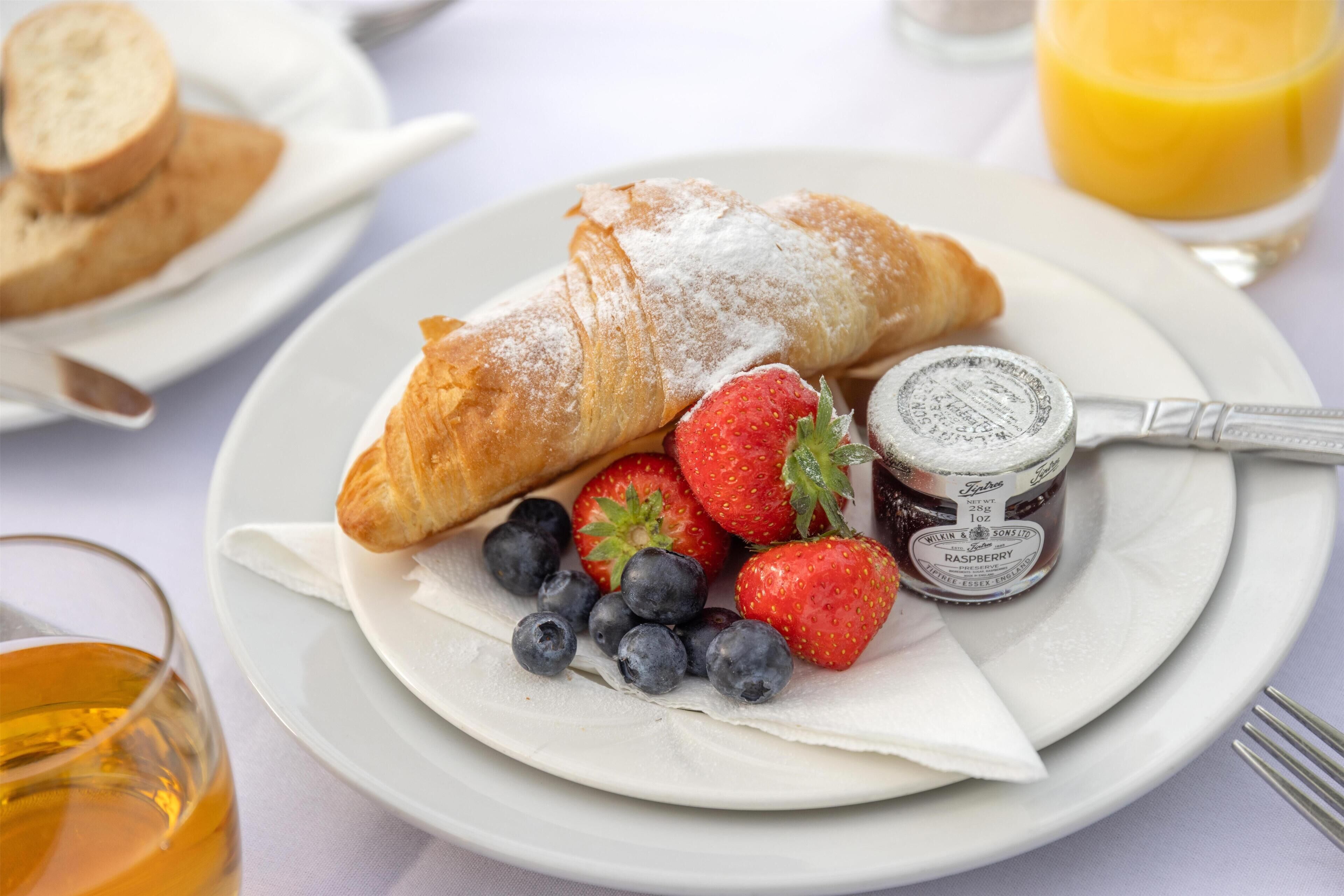 Een bord met een croissant, aardbeien, bosbessen en een pot frambozenjam.