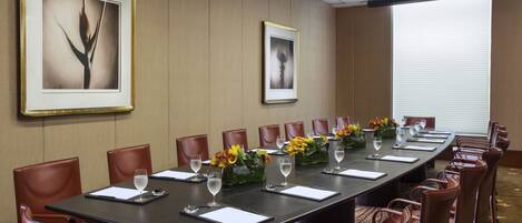 A conference room with a long table, brown leather chairs, framed artwork on the walls, and a large window with blinds.