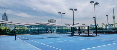 An outdoor tennis court with a net, seating area, and modern buildings in the background.