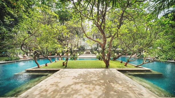 Zona de piscina con un paseo empinado, rodeada de árboles y vista despejada de un edificio al fondo.