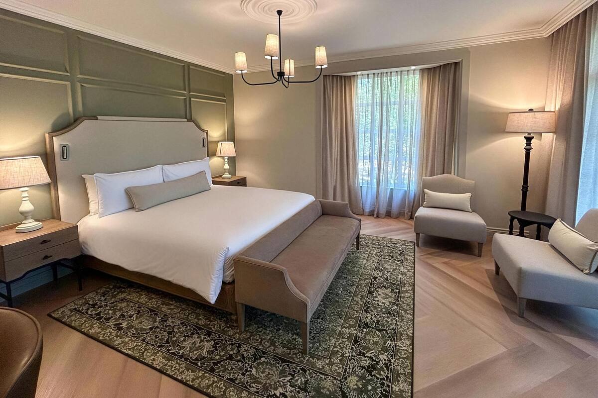 Ein Hotelzimmer mit einem großen Bett, zwei Nachttischen, einem Beistelltisch, einem Sessel, einer Stehlampe und einem Fenster mit Vorhängen.