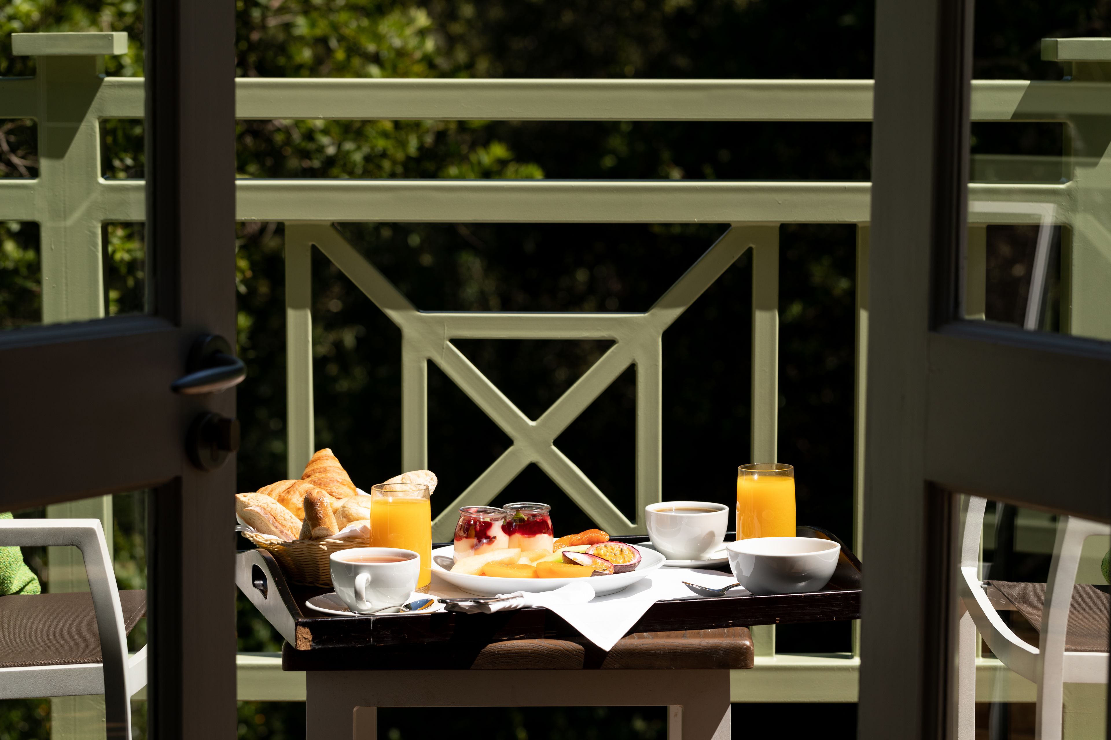 Bandeja de desayuno con cruasanes, fruta y bebidas sobre una mesa junto a una ventana.