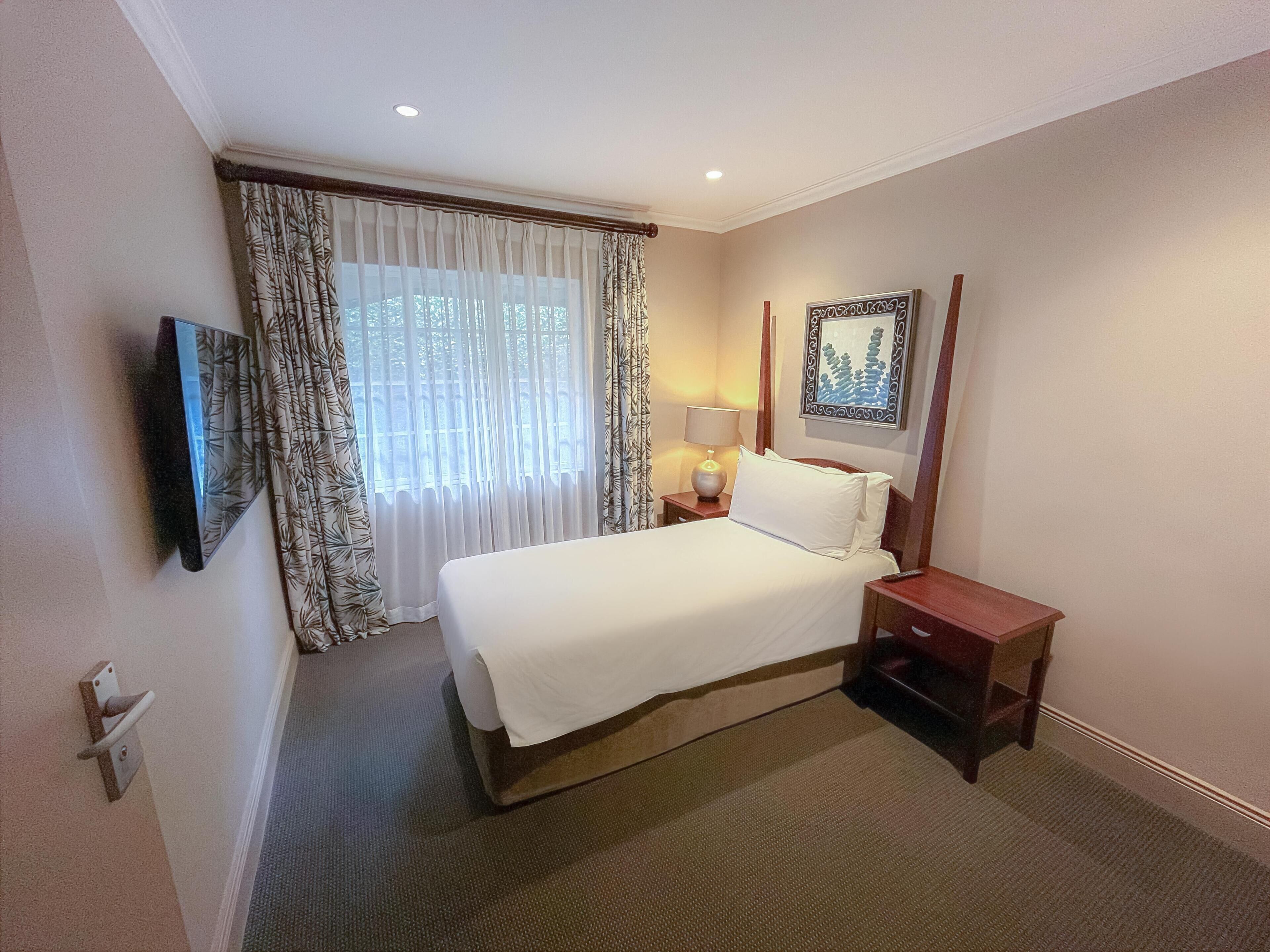 A hotel room with a bed, a bedside table with a lamp, a TV mounted on the wall, and a window with curtains.