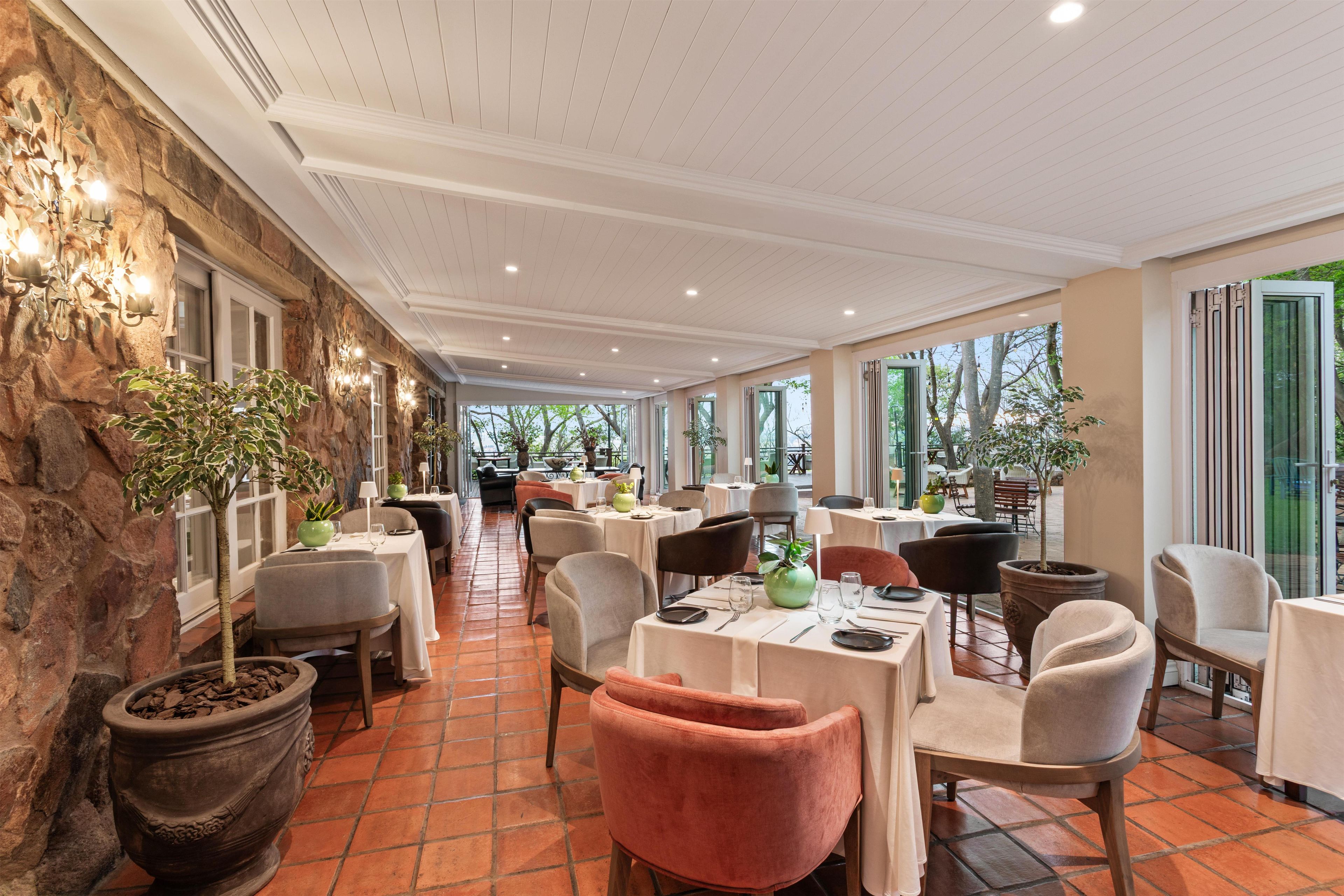 A restaurant with tables and chairs arranged for dining, a stone wall on one side, and large windows allowing natural light.