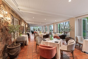 Un restaurante con mesas y sillas dispuestas para comer, una pared de piedra en un lateral y amplias ventanas que dejan entrar luz natural.