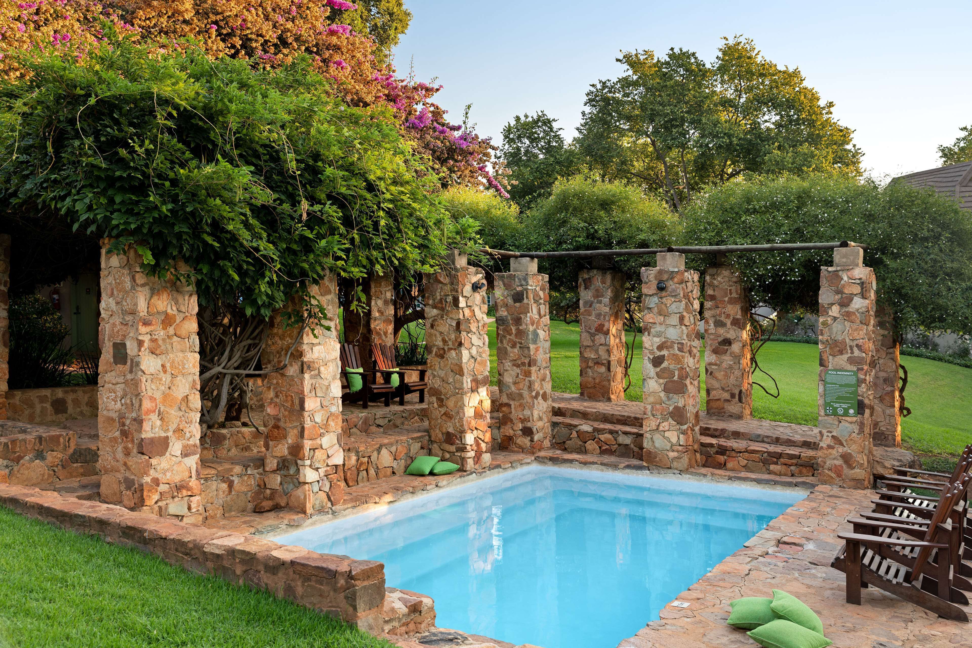 Una piscina de pedra amb una pèrgola, cadires verdes i una zona enjardinada.