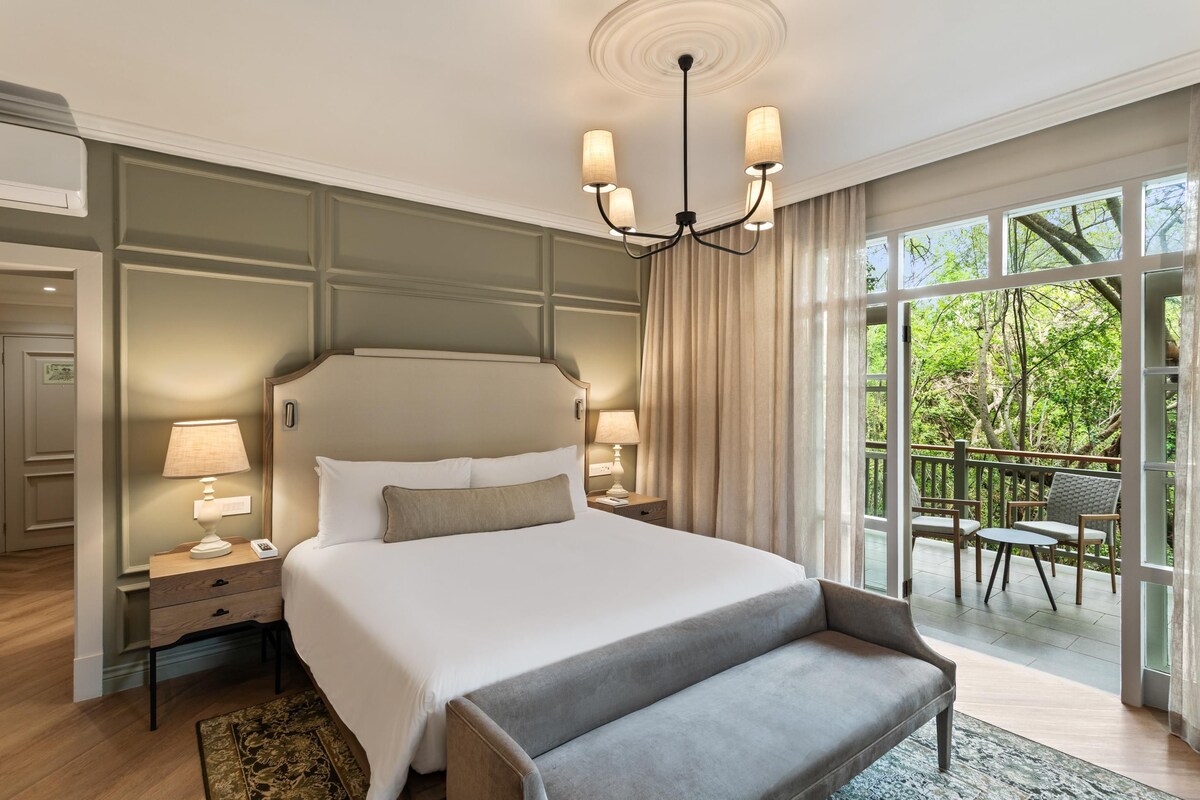 A hotel room with a large bed, two bedside tables, a chandelier, a balcony with a view of trees, and a seating area.