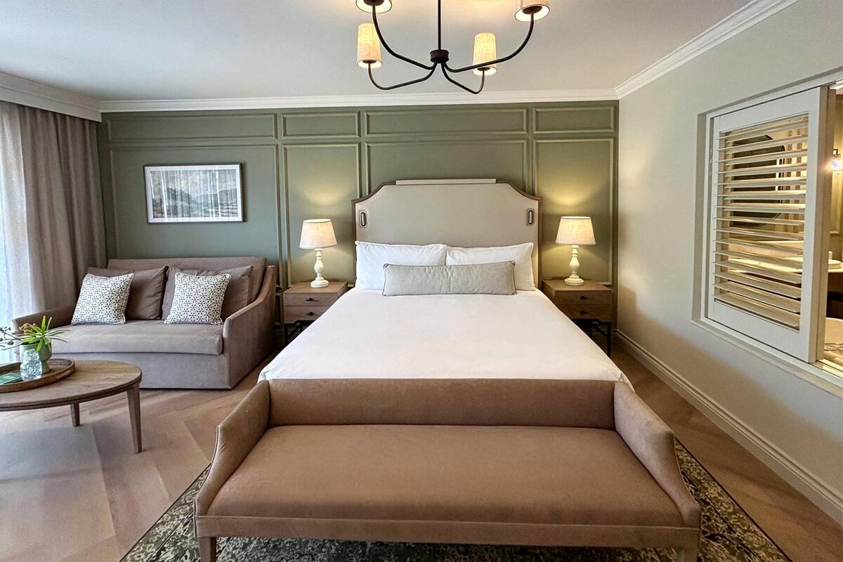 Ein Hotelzimmer mit einem großen Bett, einer Couch, einer Bank, zwei Nachttischen mit Lampen, einem Chandelier und einem Fenster mit Jalousien, sowie einem gerahmten Bild an der Wand.