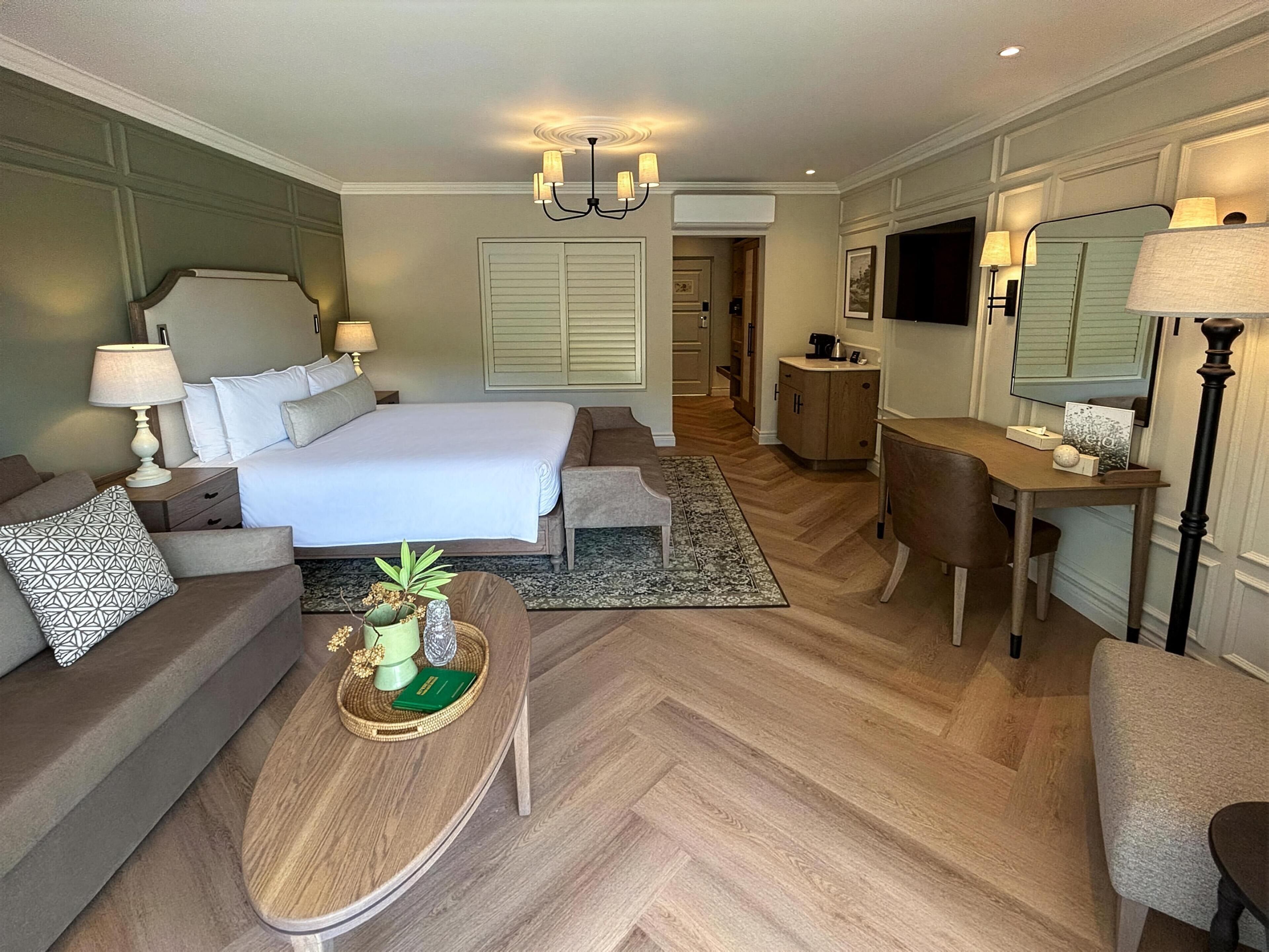 Habitación de hotel con una cama grande, zona de estar con sofá y sillón, escritorio con silla, una mesita con objetos decorativos y un espejo grande.