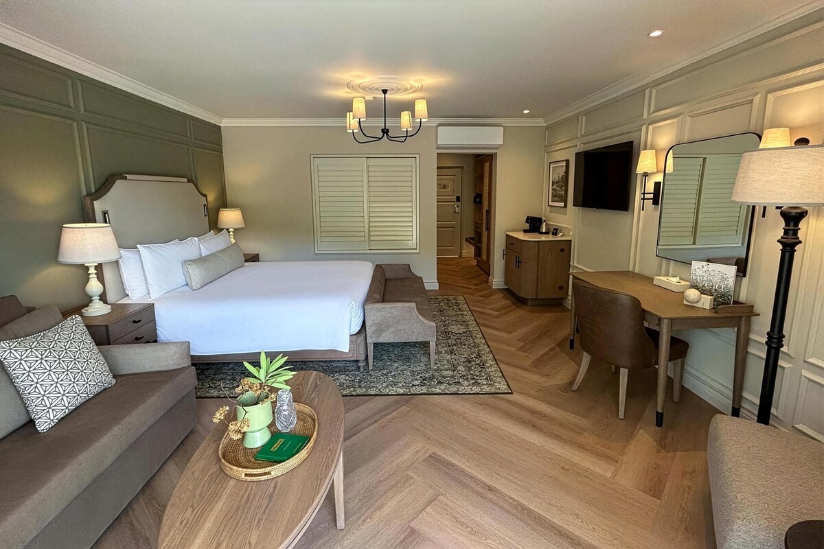 A hotel room with a large bed, a sitting area with a sofa and armchair, a desk with a chair, a small table with decorative items, and a large mirror.