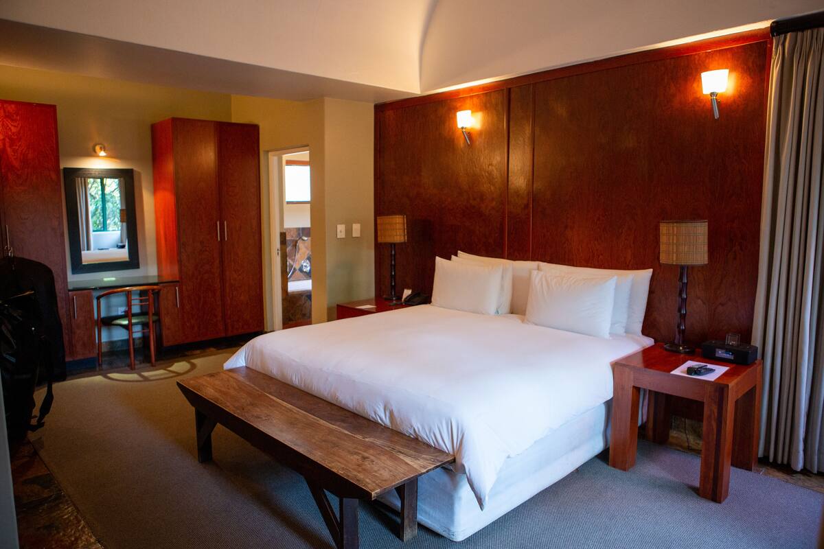 A hotel room with a large bed, wooden headboard, two bedside tables with lamps, a bench, a desk with a chair, and a mirror.
