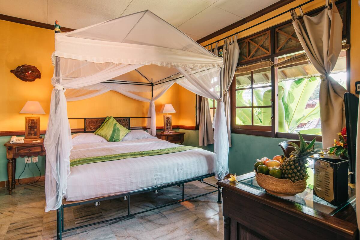 A room with a four-poster bed, a nightstand with a lamp, a window with curtains, and a fruit basket on a table.