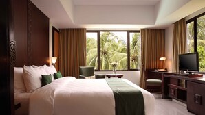A hotel room with a large bed, a desk with a TV, and a view of palm trees through the windows. - Aryaduta Lippo Village (Curug)