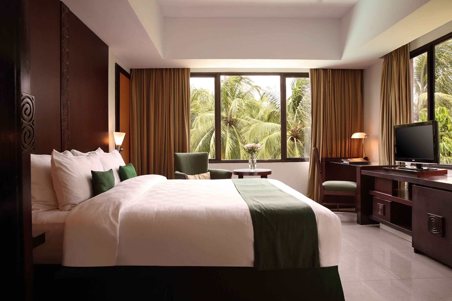 A hotel room with a bed, desk, chair, TV, and large windows with curtains.