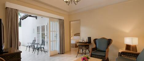 A spacious living room with a sofa, armchair, and a coffee table with a vase of flowers. There is a dining area with chairs and a table, and a bedroom visible through an open door.