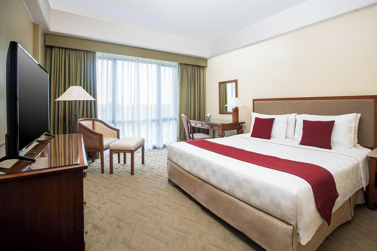 A hotel room with a large bed, a desk, a chair, a television, and a window with curtains.