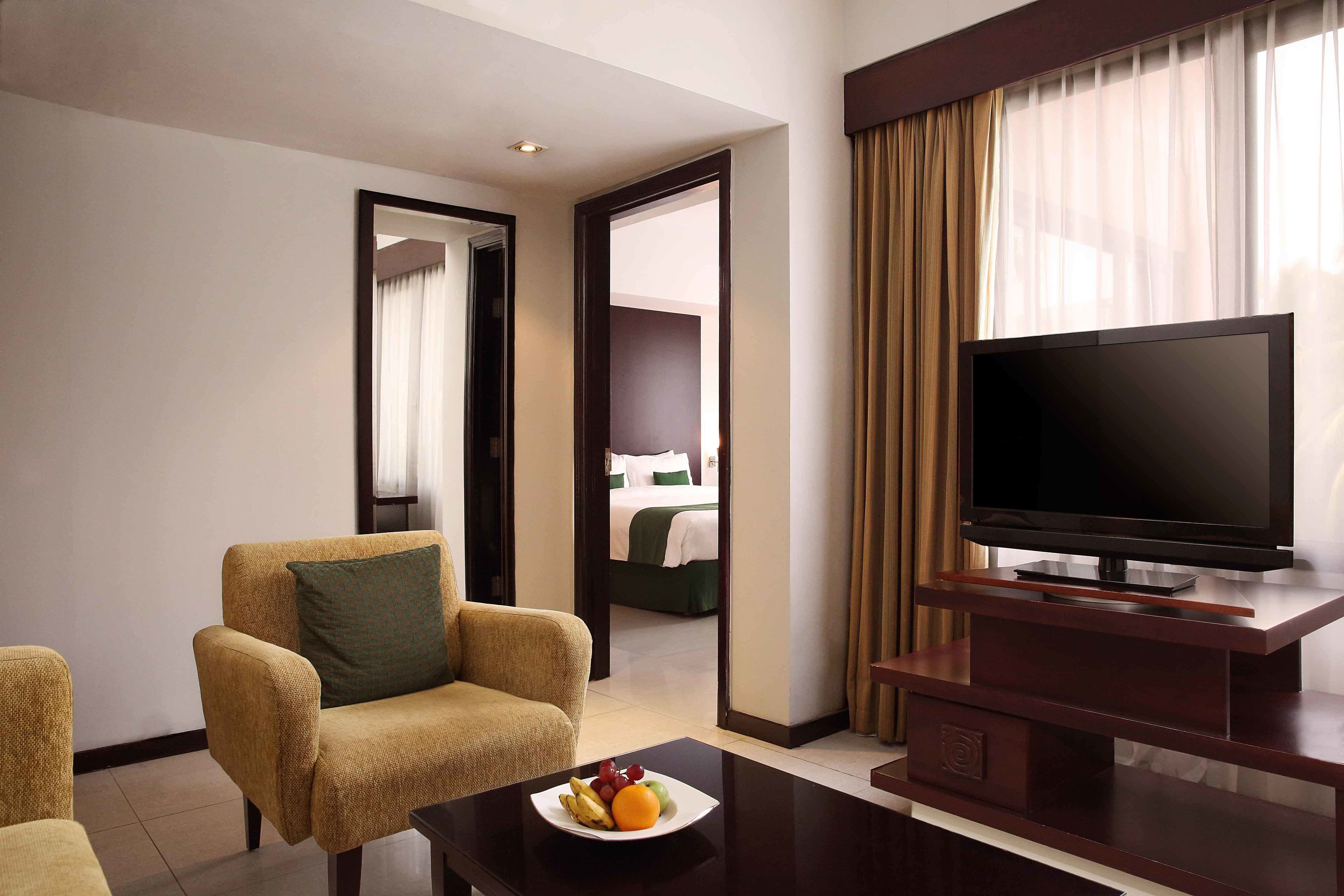 Habitación de hotel con una televisión sobre un mueble de madera, un sofá con un cojín verde y una mesa con fruta.