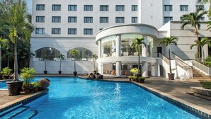 A hotel with a swimming pool, palm trees, and a modern building with large windows and a curved glass structure. - Aryaduta Lippo Village (Curug)