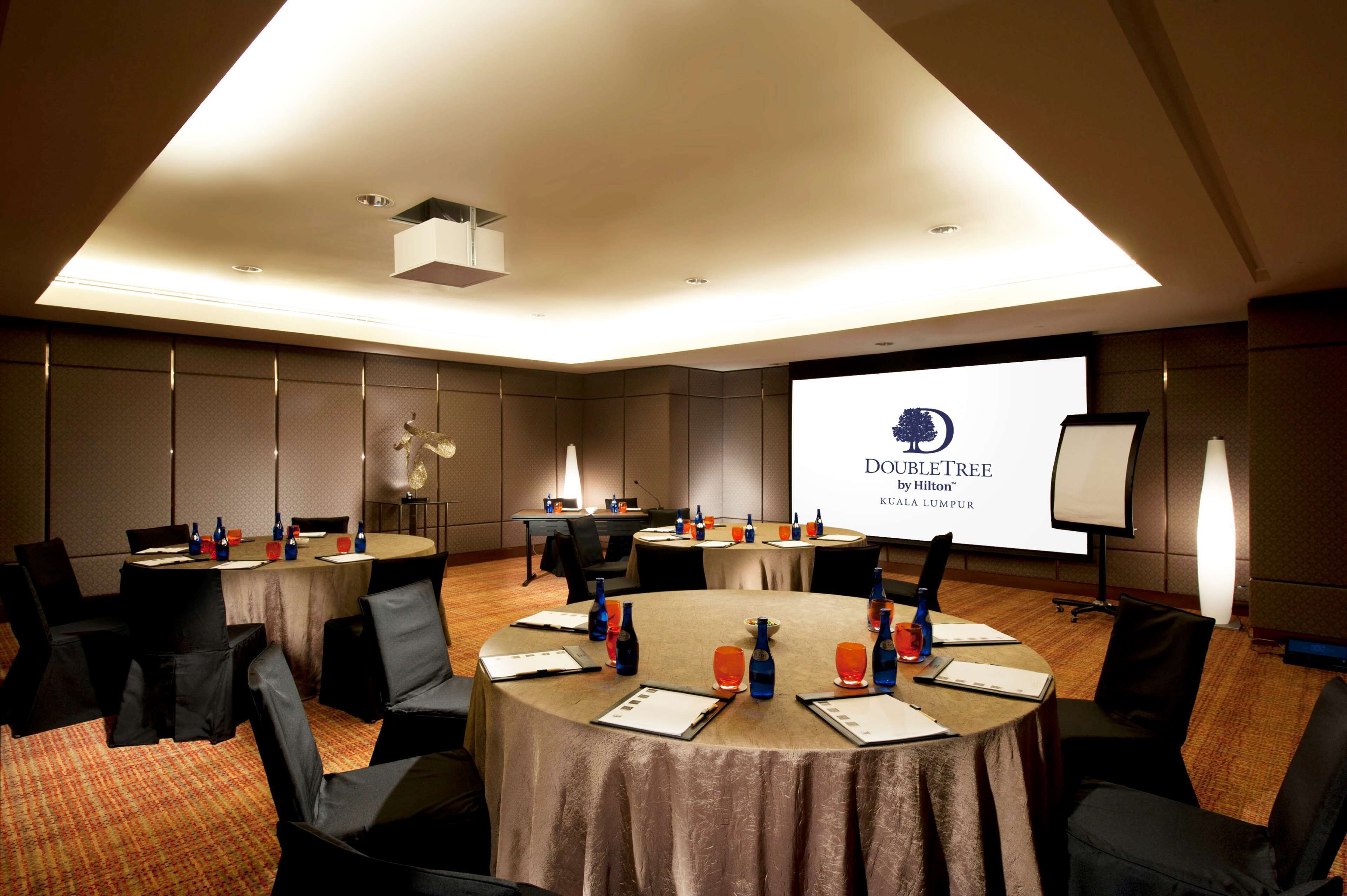 A conference room with round tables, chairs, and a projector screen displaying 'Doubletree Kuala Lumpur'.