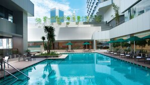 A modern indoor swimming pool with lounge chairs and umbrellas, surrounded by high-rise buildings and greenery.