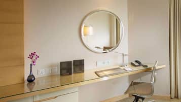 Habitación de hotel con escritorio de vidrio, espejo redondo, florero con flores, teléfono y una silla.