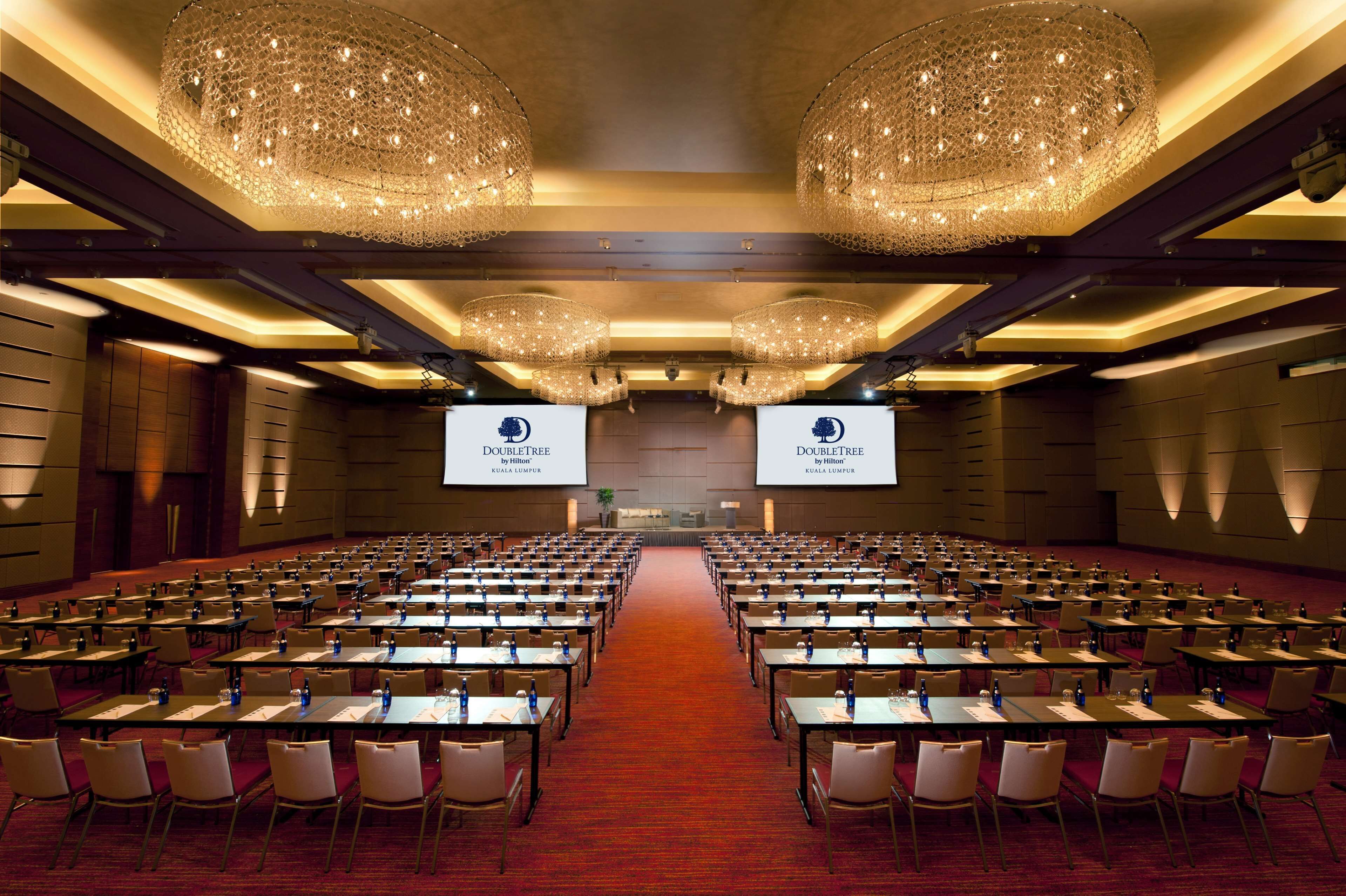 Un salón de conferencias con filas de mesas y sillas, grandes candelabros y dos pantallas que muestran el logotipo de DoubleTree Kuala Lumpur.