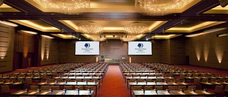 A conference room with rows of tables and chairs, large chandeliers, and two screens displaying the DoubleTree Kuala Lumpur logo.