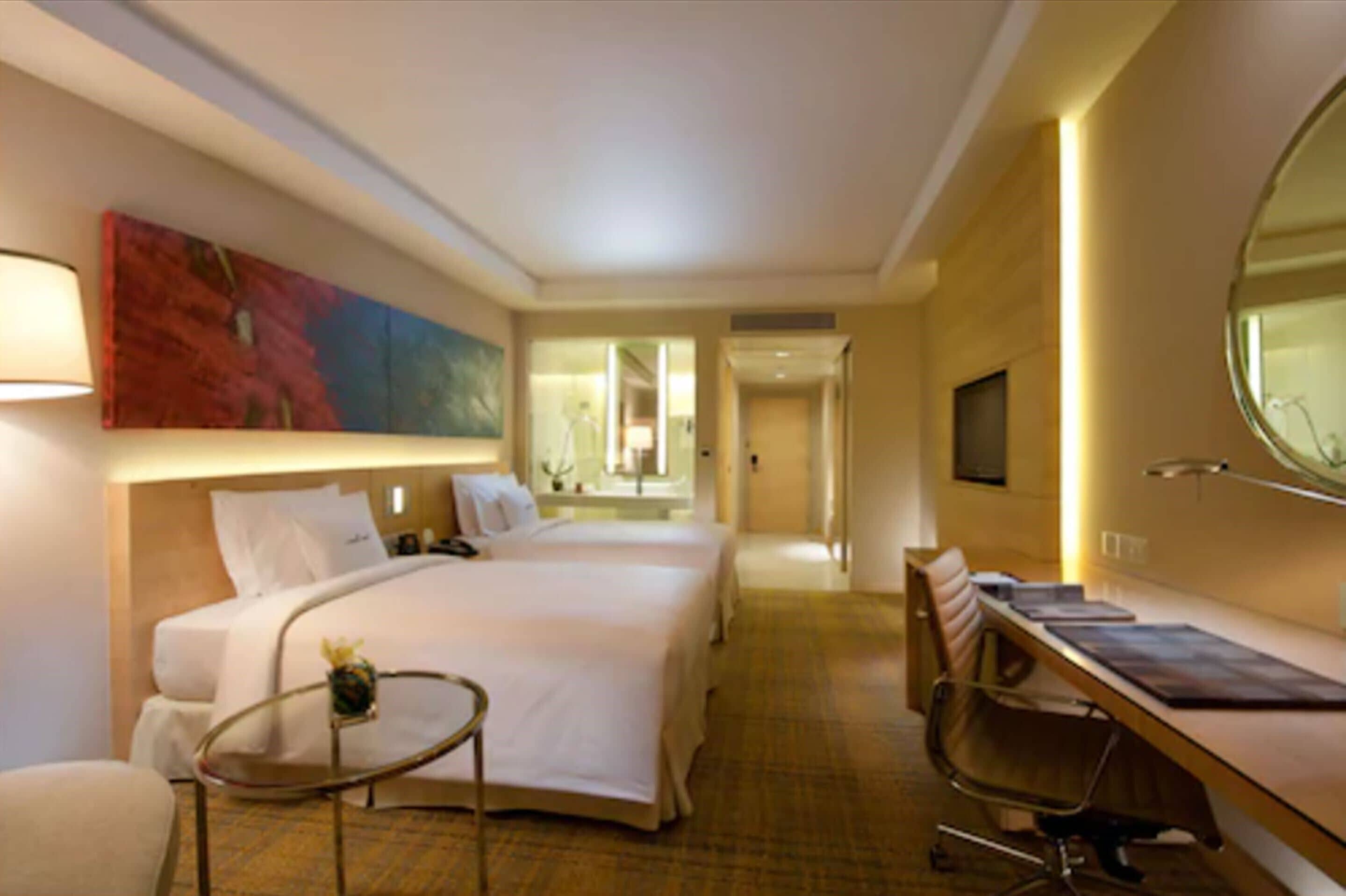 A hotel room with two beds, a desk, a chair, a small table with a vase, and a large painting on the wall.