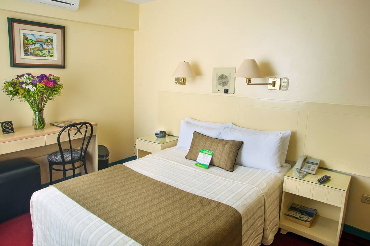 A hotel room with a bed, bedside tables, a desk, a chair, a vase of flowers, and wall-mounted lamps.