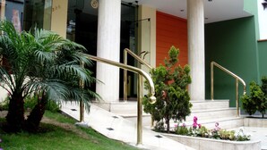 Un bâtiment moderne doté de grandes colonnes, d’un escalier orné de rampes dorées et d’un petit jardin planté de palmiers et de fleurs.