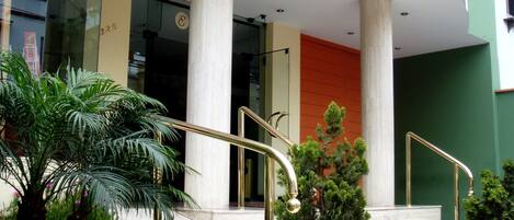Un edificio moderno con grandes columnas, una escalera con barandales dorados y un pequeño jardín con palmeras y flores.