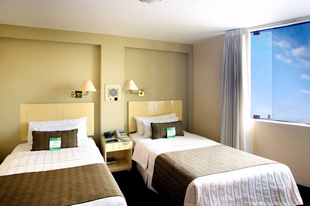 A hotel room with two beds, a nightstand, a lamp, a phone, and a window with curtains.