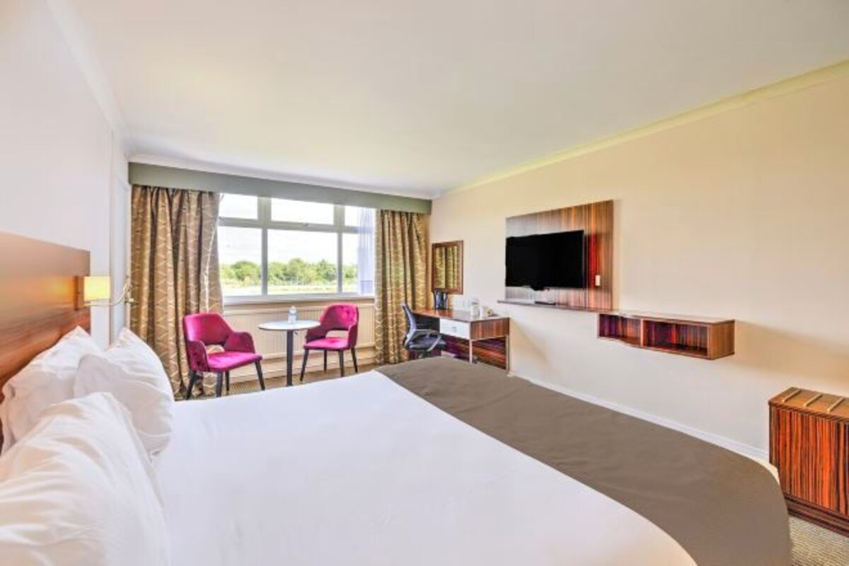 Habitación de hotel con una cama grande, un escritorio, dos sillas, un televisor y una ventana con cortinas.