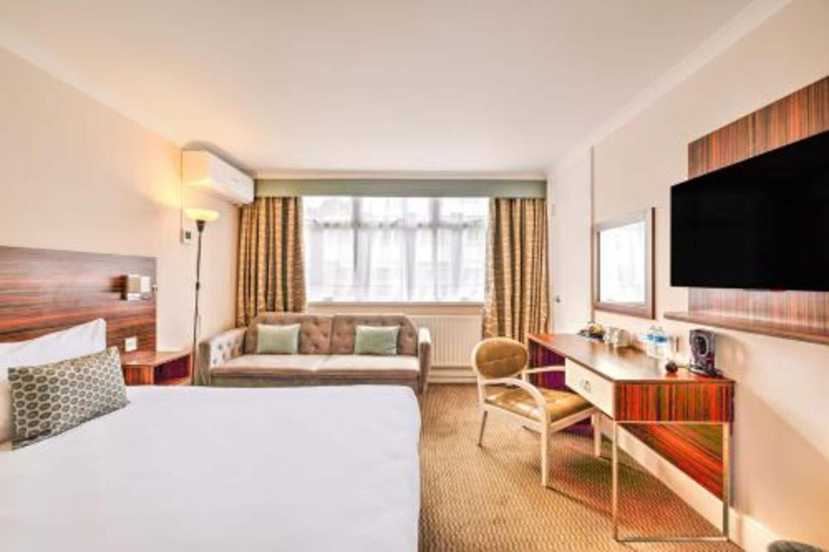 A hotel room with a large bed, a desk with a chair, a TV mounted on the wall, and a window with curtains.