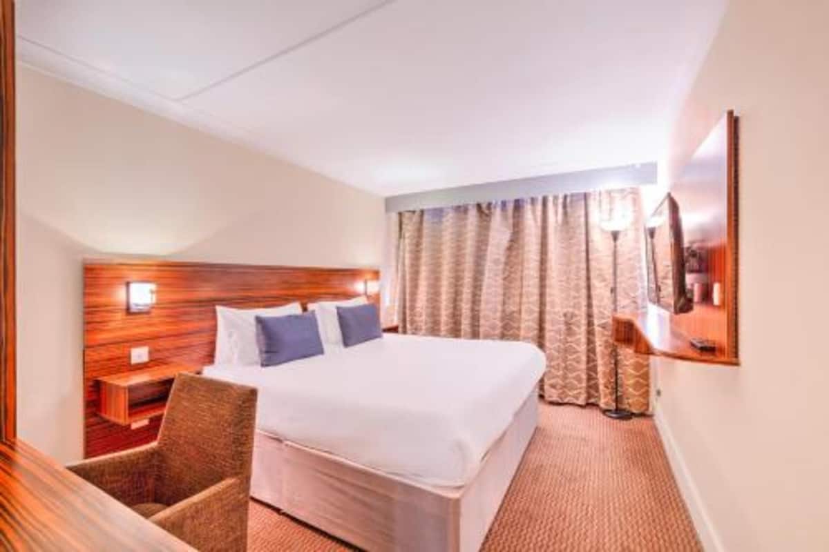 Habitación de hotel con una cama grande, cabecera de madera, mesita de noche, una silla y un televisor en la pared.
