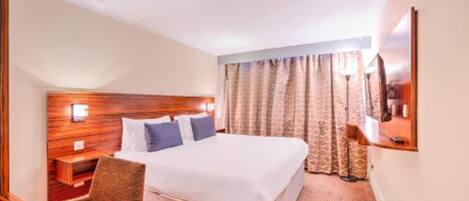 Ein Hotelzimmer mit einem großen Bett, einem Holzkopfteil, einem Nachttisch, einem Sessel und einem an der Wand montierten Fernseher.