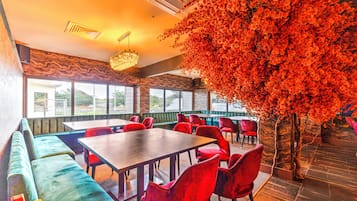A restaurant with a large table, red chairs, a green cushioned bench, and a tree with red flowers.