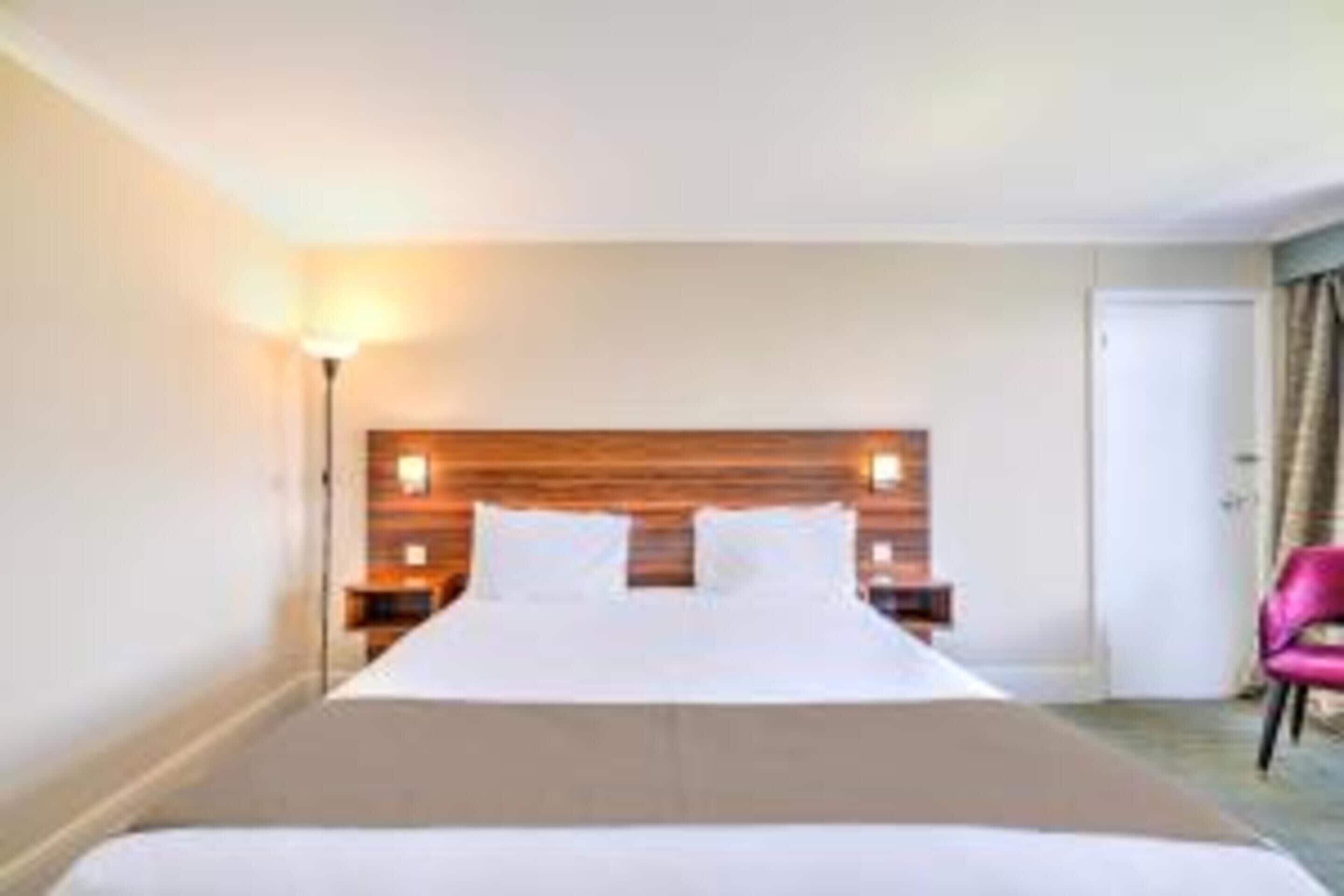 A hotel room with a large bed, a wooden headboard, a nightstand, a lamp, and a pink chair.