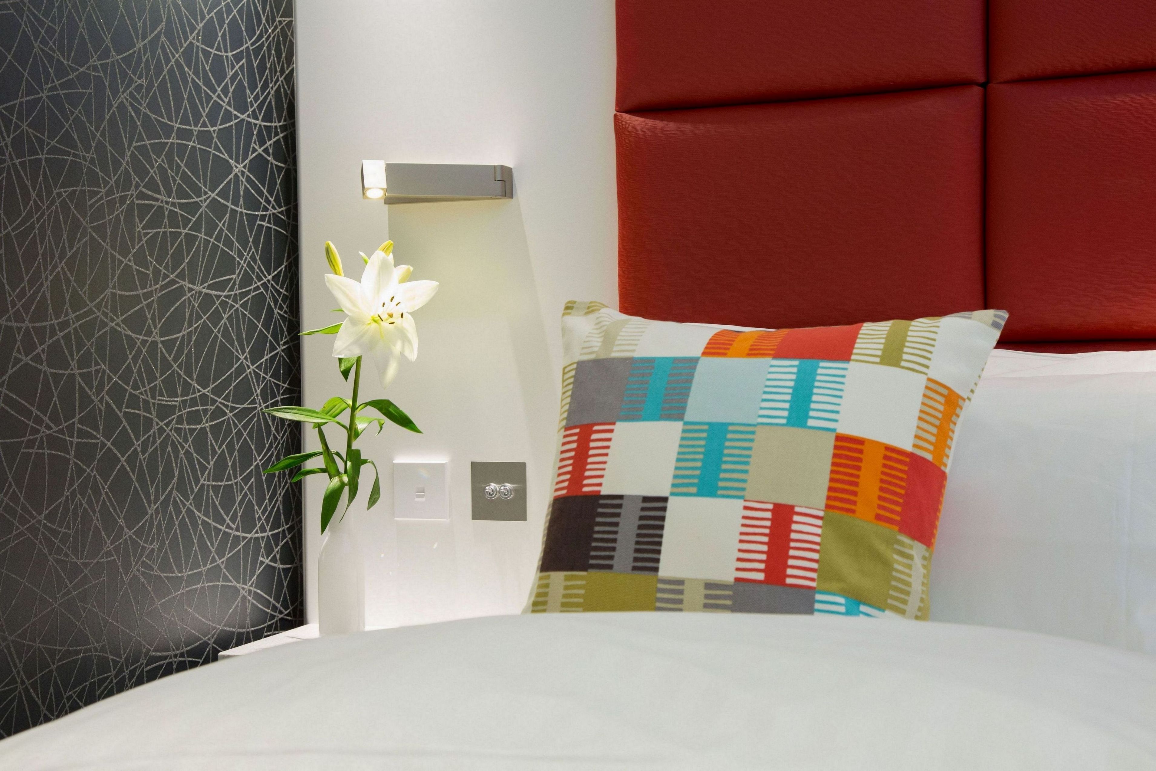 A modern hotel room with a bed, a colorful pillow, a vase with a white flower, and a wall with a geometric pattern.