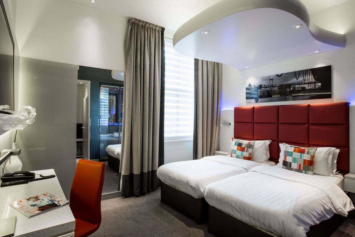 A modern hotel room with a large bed, a desk with a chair, a mirror, and a window with curtains.