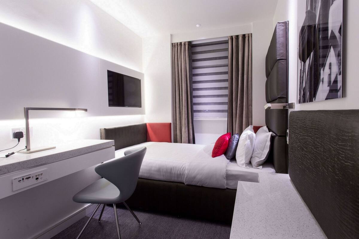 A modern hotel room with a bed, a desk, a chair, a TV, and a window with curtains.